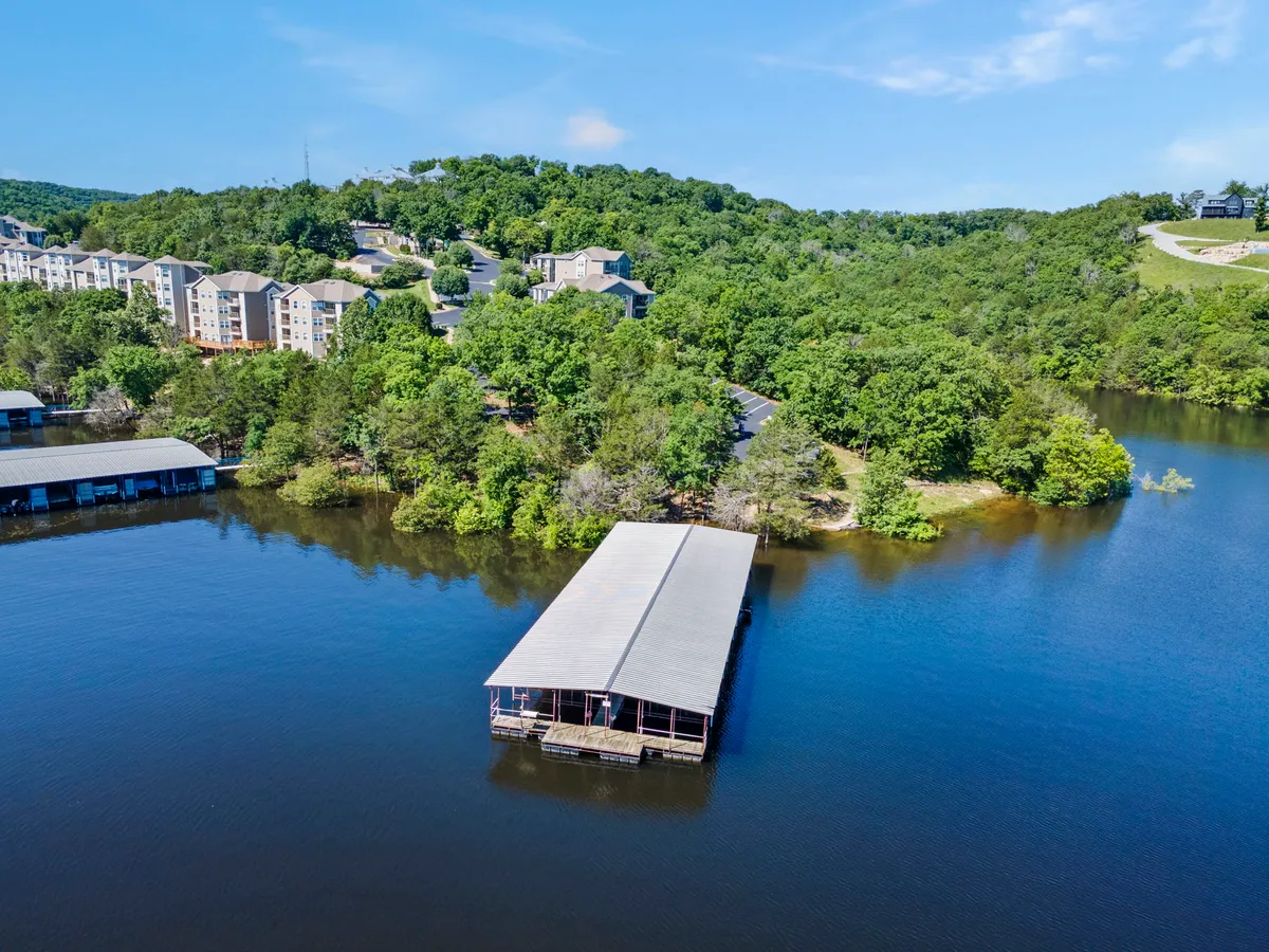 A peaceful dock extends into the water, surrounded by lush greenery, with residential apartments visible on the hill beyond. The area invites boating, fishing, and picnicking right along the shore, evoking a sense of community and lakeside enjoyment.