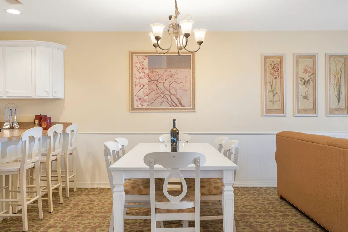 A cozy, warmly decorated interior featuring a central dining table under a chandelier, with a comfortable couch and bar stools nearby. The space blends dining and lounging areas, ideal for family meals, celebrations, and casual gatherings.