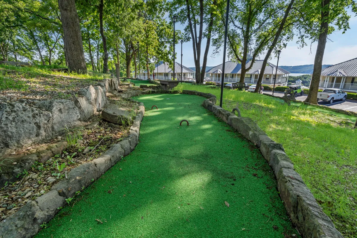 A vibrant mini-golf course with lush green landscaping and natural stone borders sits beneath shady trees. The scene shows families and children engaged in a friendly competition, clearly enjoying themselves in a playful, picturesque setting ideal for all ages.