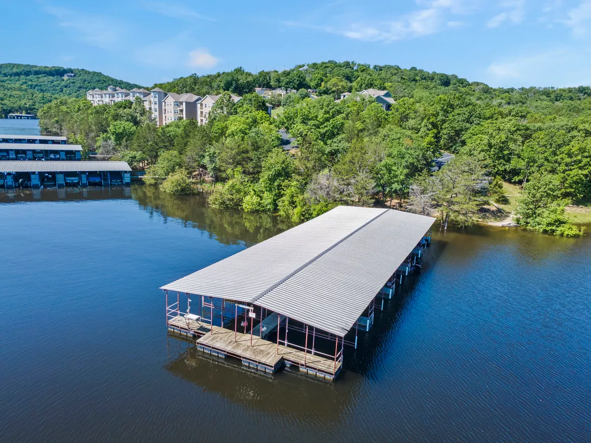This image features a tranquil boat dock extending over the calm water, surrounded by lush greenery with apartments visible in the background. The scene captures the easy lake access and serene environment guests can enjoy right outside the condo.
