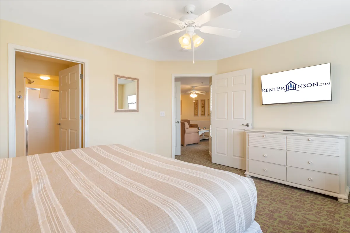 A stylish bedroom featuring a king bed with striped bedding, a wall-mounted TV, a dressing mirror, and sunlight streaming in from the side. The room feels open and tranquil, with doorways leading to other private spaces, suggesting ensuite access.