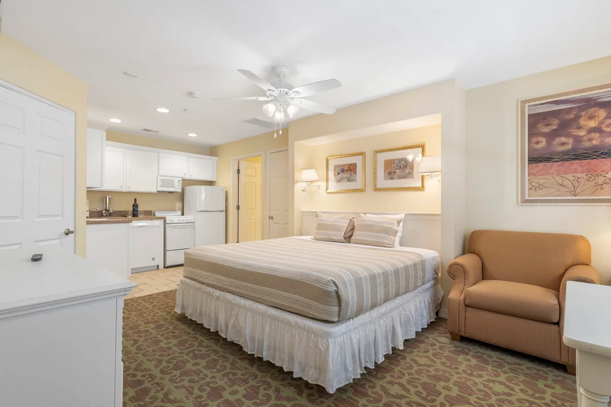 A bedroom with a king bed, armchair, and a compact kitchenette in the corner, decorated with artwork and natural light. The room exudes a sense of convenience and comfort, perfect for unwinding.