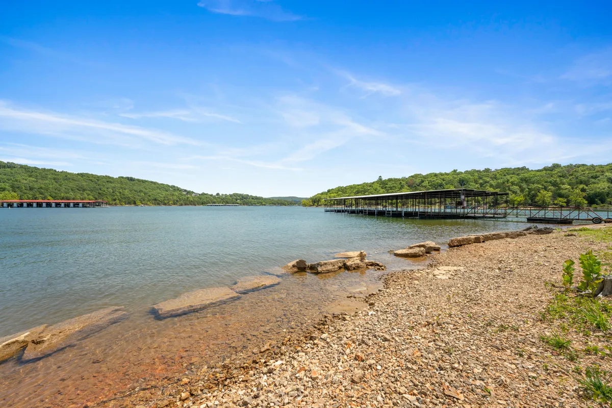 A serene lakeside view showing calm water, a charming boathouse, distant hills, and a rocky shoreline. The scene invites quiet reflection, fishing, or nature walks, with soft light suggesting early morning or twilight hours.
