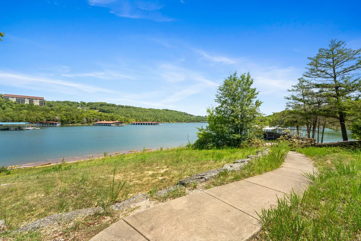 A broad perspective of the lake, with a winding pathway along the shore, lush greenery, and distant hills. The scene is calm and inviting, suggesting opportunities for walking, wildlife observation, and lakeside picnics while taking in the Ozark sunsets.