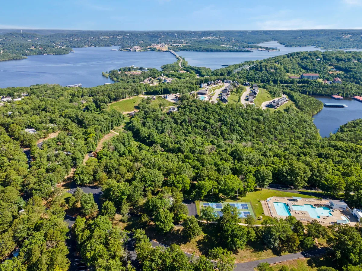 A vibrant aerial view of the resort grounds, featuring a sparkling swimming pool, visible tennis courts, and the serene lake surrounded by lush greenery. Families can be seen swimming and gathering around the pool, while others enjoy recreational activities in the open spaces.