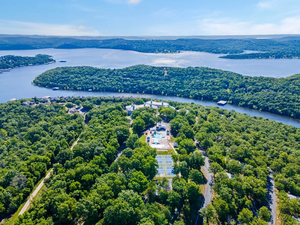 A broad exterior shot features a sparkling outdoor swimming pool set against an expansive lake, with tennis courts and lush green trees surrounding the resort area. The sun is low, suggesting a golden-hour ambiance perfect for evening activities.