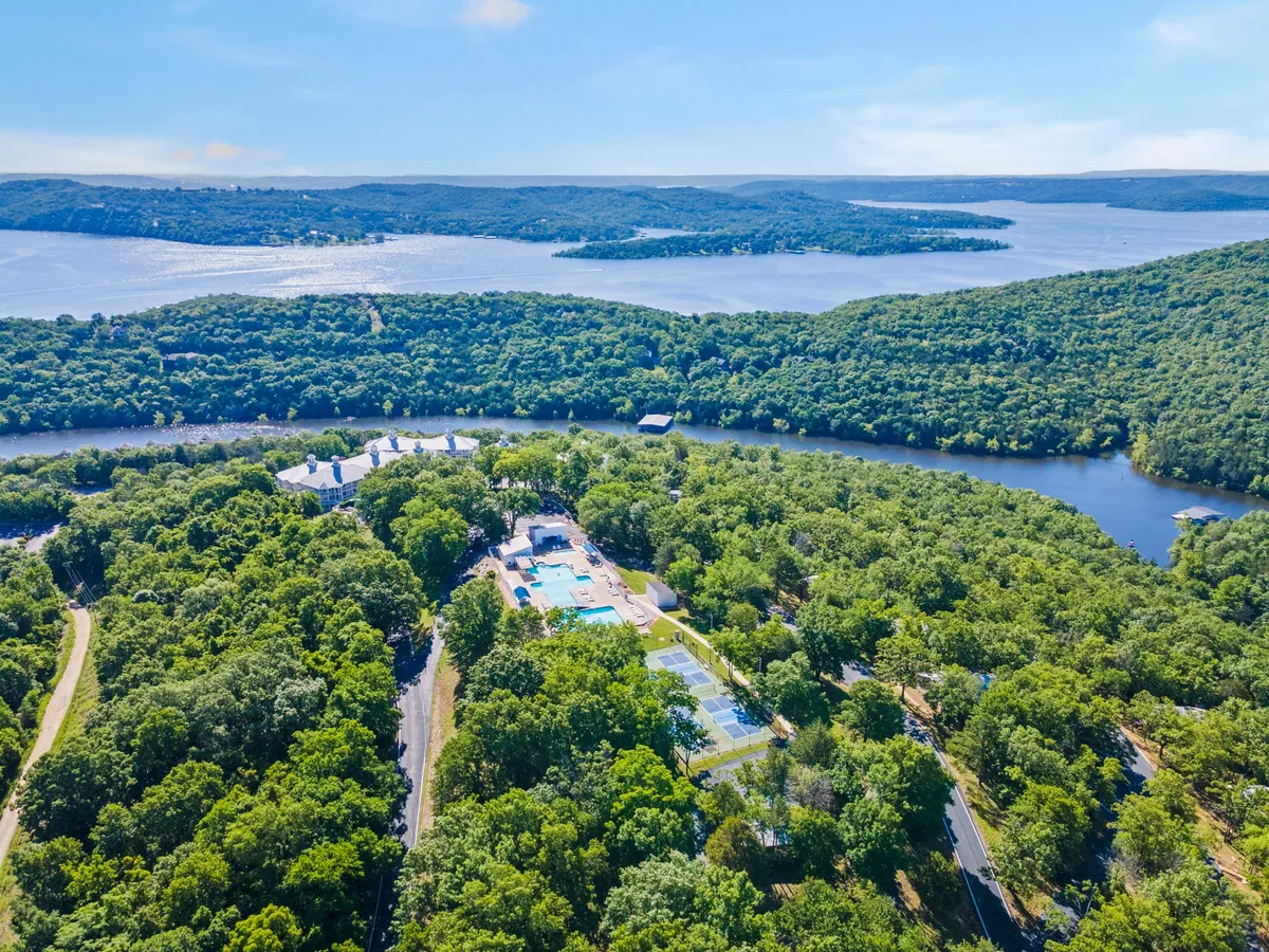 An inviting aerial view of a resort area with a shimmering swimming pool, tennis courts, dense forests, and a picturesque river bend. The landscape glows under the sun, evoking a sense of tranquility and opportunity for outdoor adventure.